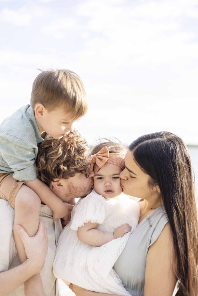 A beautiful photo of a  mum and dad kissing their little girl as big brother watches on sitting o n dad's shoulders | Brisbane Family Photoshoot | Sharon Joseph Photography