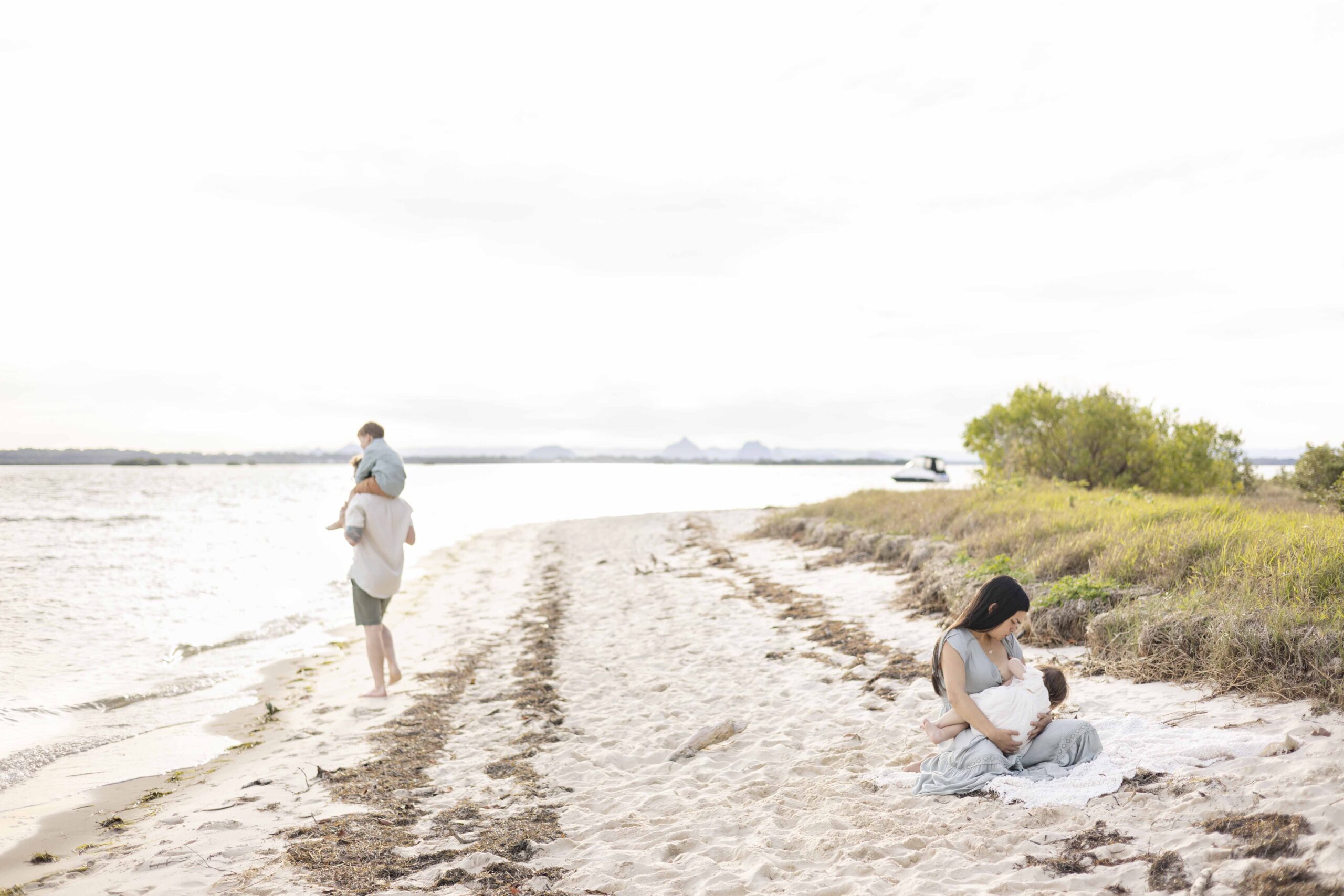 Brisbane lifestyle family photos. The Mum is breastfeeding her little baby girl. The little boy is sitting on his dad's shoulders and looking out to the sea. By Sharon Joseph Photography, Brisbane Family Photographer.