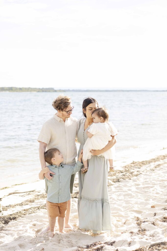 Outdoor family photos in North Brisbane | Family on the beach. The mum gently kisses her little girl's forehead as the dad and brother lovingly watch on | Sharon Joseph Photography