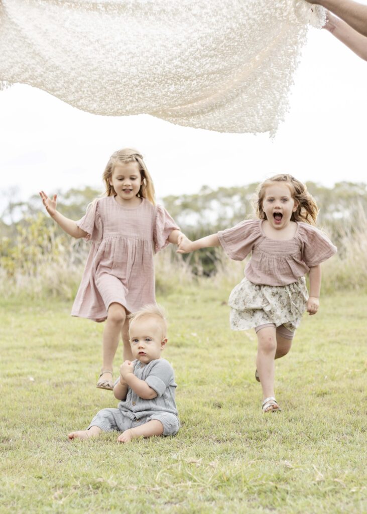 Two little girls joyfully run under a blanket "parachute" as their little brother watches on. Brisbane Family Photography by Sharon Joseph