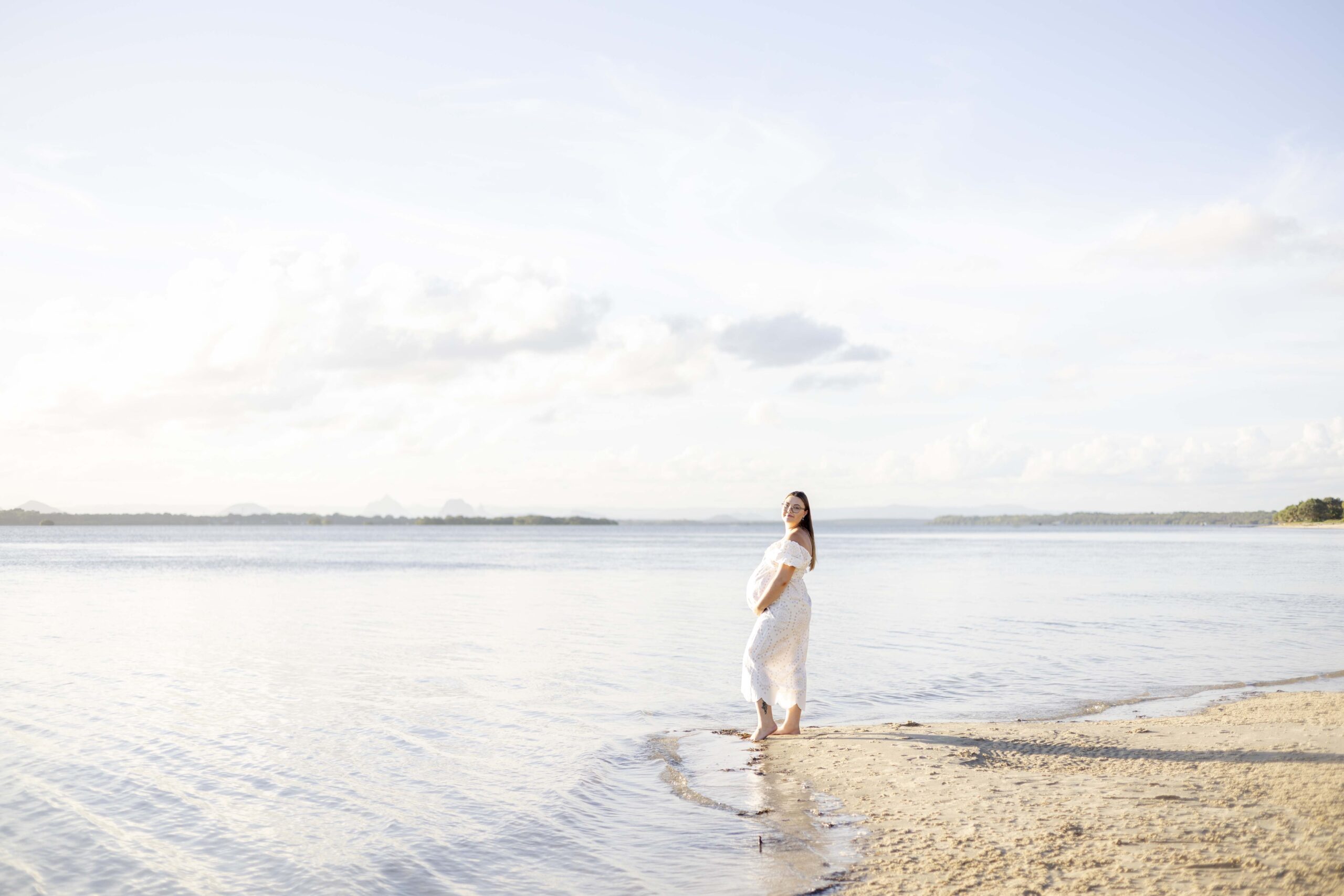 A beautiful expectant mum standing at the water's edge on the beach in Brisbane