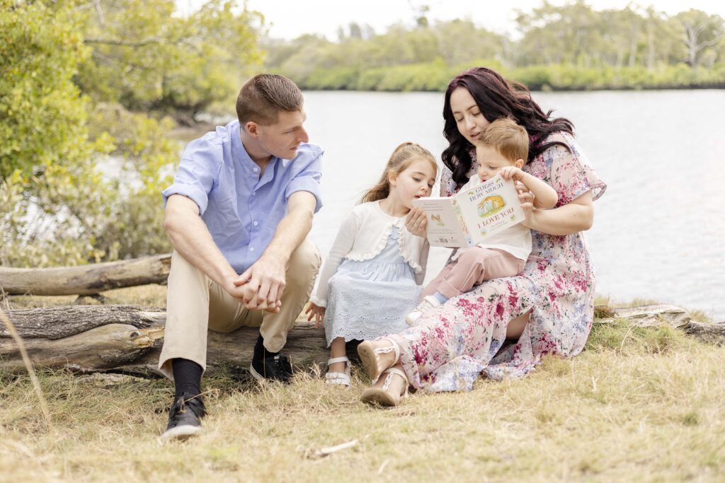 Family reading the book "Guess how much I love you" while sitting on a log by the river in Brisbane, on a cloudy day.