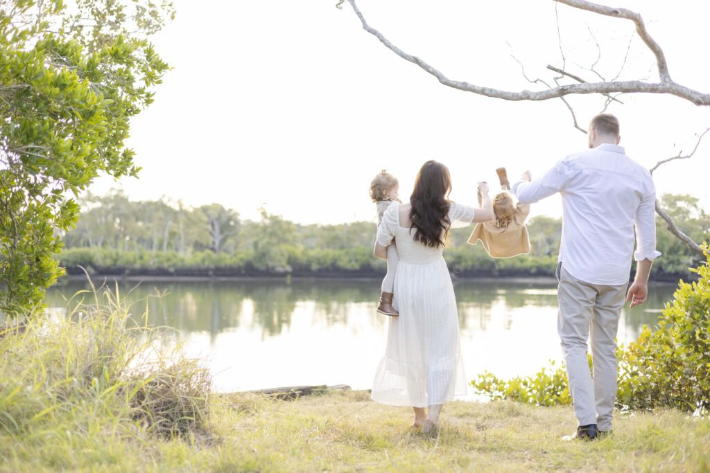 Family of four near a river bank on a sunny afternoon. The mum and dad are swinging their little girl.