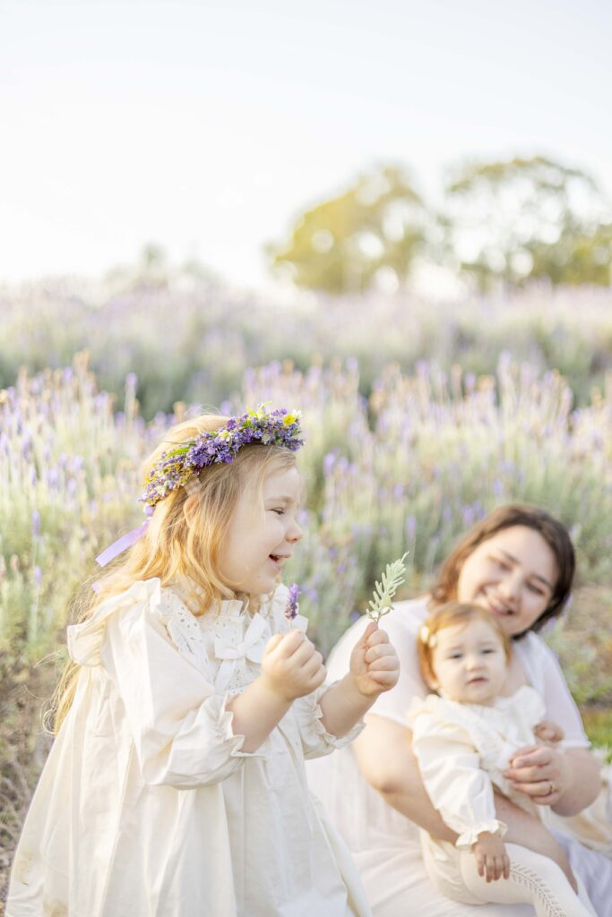 Little girl happily holding a lavender flower as her mum and baby sister watch on with joy, at their relaxed family photo session in Brisbane