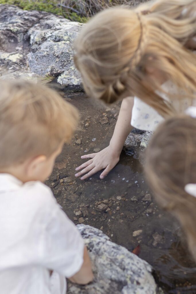Girl dipping her hand in a puddle as her siblings watch on.