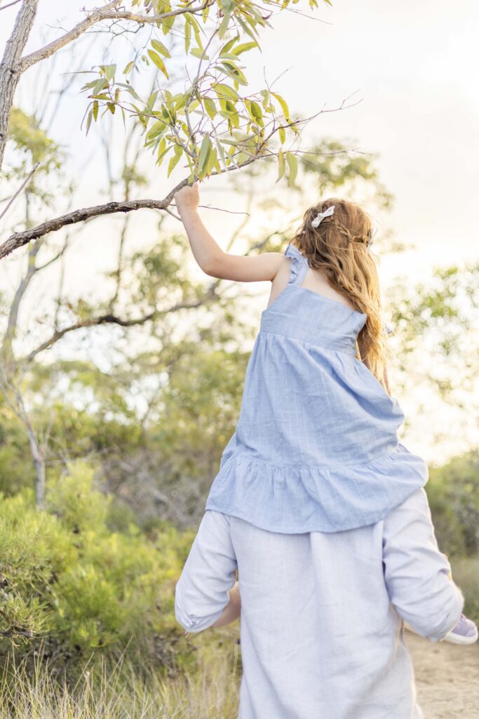 Girl on dad's shoulder, reaching for a tree branch during their family bushwalk near Brisbane/Sunshine Coast