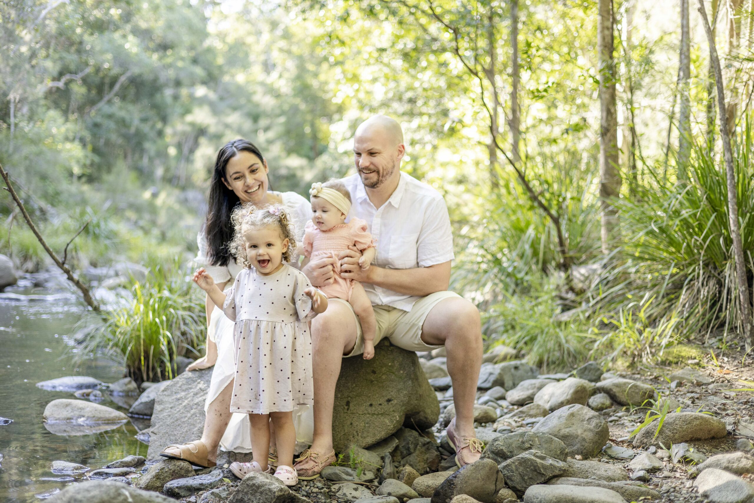 happy photo of little girl giving a big cheeky smile while her parents look at her, at their family photo session at a creek near Brisbane