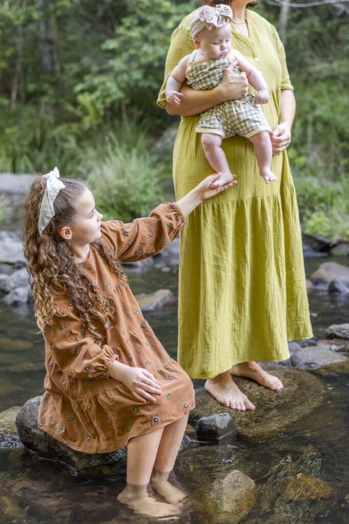Big sister gently stroking baby sister's feet, at their relaxed family photography session at a creek near Brisbane
