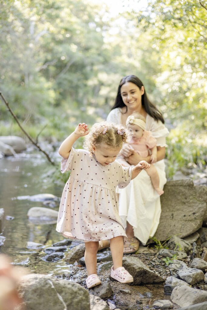 Mum and baby sister watch on as toddler carefully walks over rocks near the creek, at their relaxed family photography session in Brisbane.